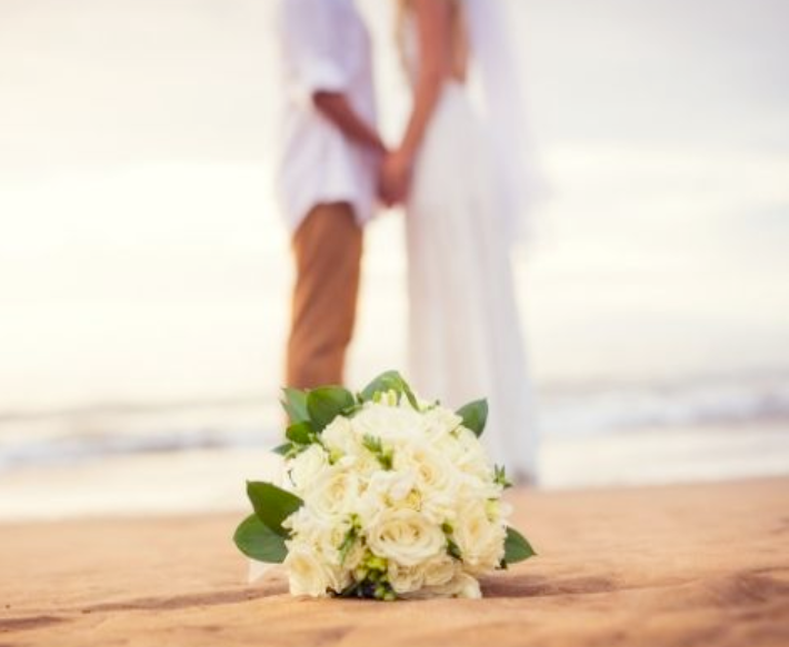 A man and woman holding hands and embracing on beach with bouquet of white flowers in front of them near Ocean City Fontainebleau Resort in Ocean City, MD